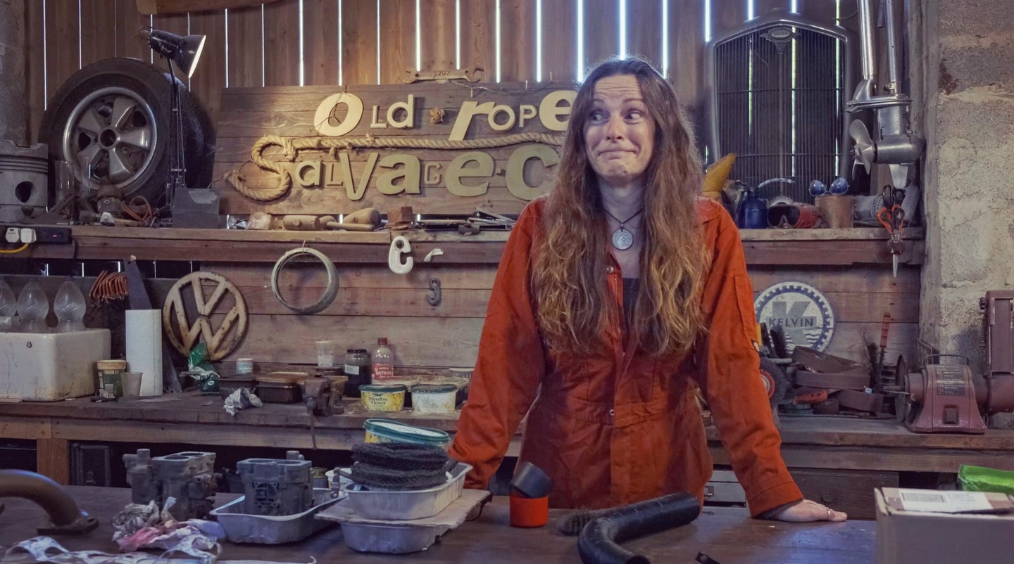 Woman in orange overalls standing behind a workbench in a workshop with a sign behind saying old rope salvage