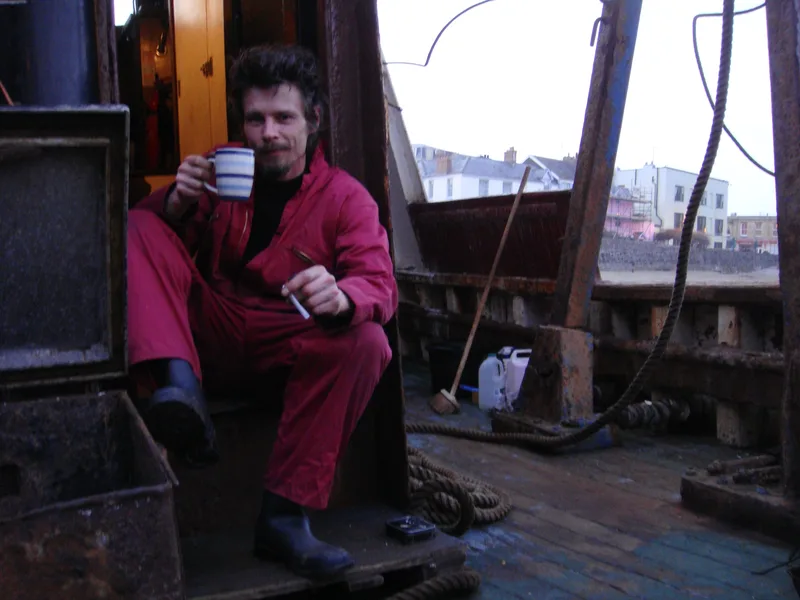 Man in red overalls sitting on the deck of a fishing trawler drinking tea