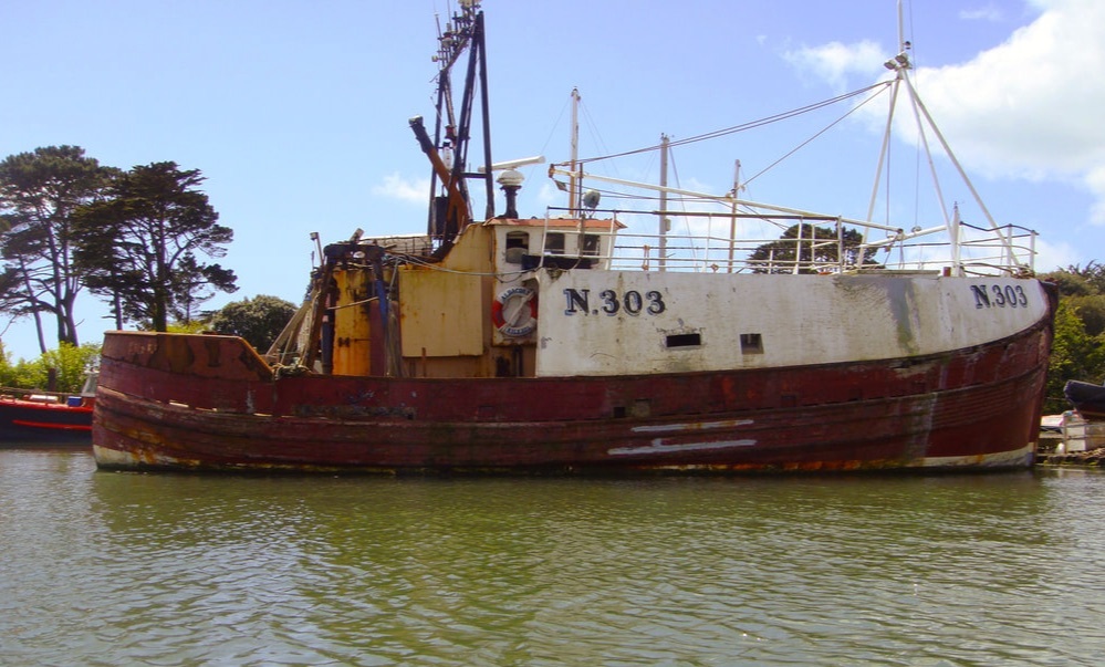 Old red and white wooden Fishing Trawler Albacore N303 moored on tranquil water with blue sky and trees behind