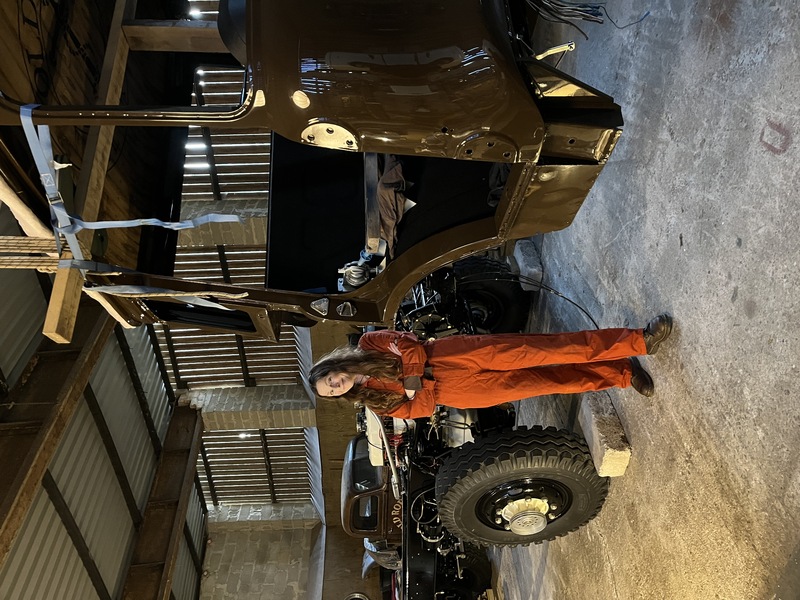 Woman in orange overalls standing next to the separated cab and chassis of a restored classic Bedford truck